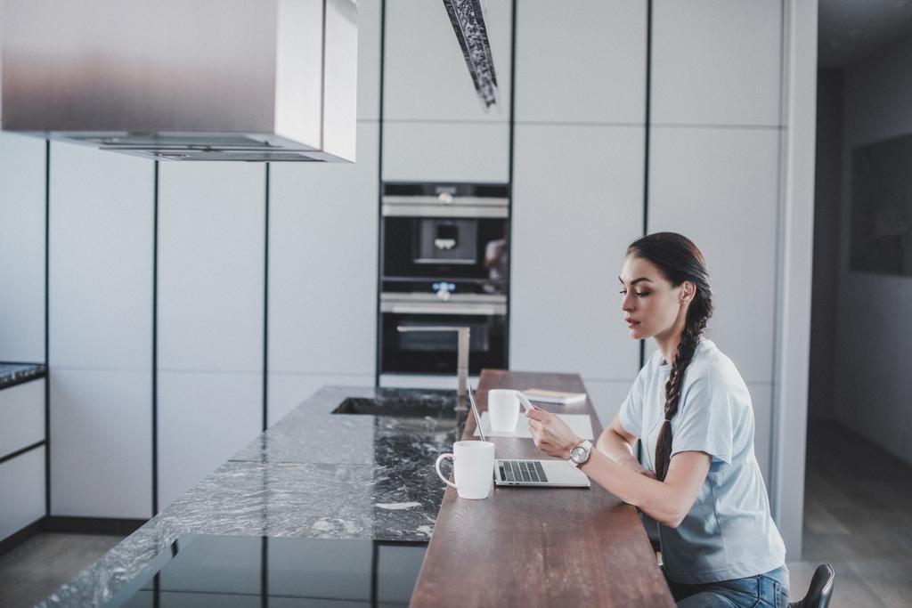 vue latérale de belle pigiste travaillant à l'ordinateur portable et tenant smartphone dans la cuisine
 - Photo, image