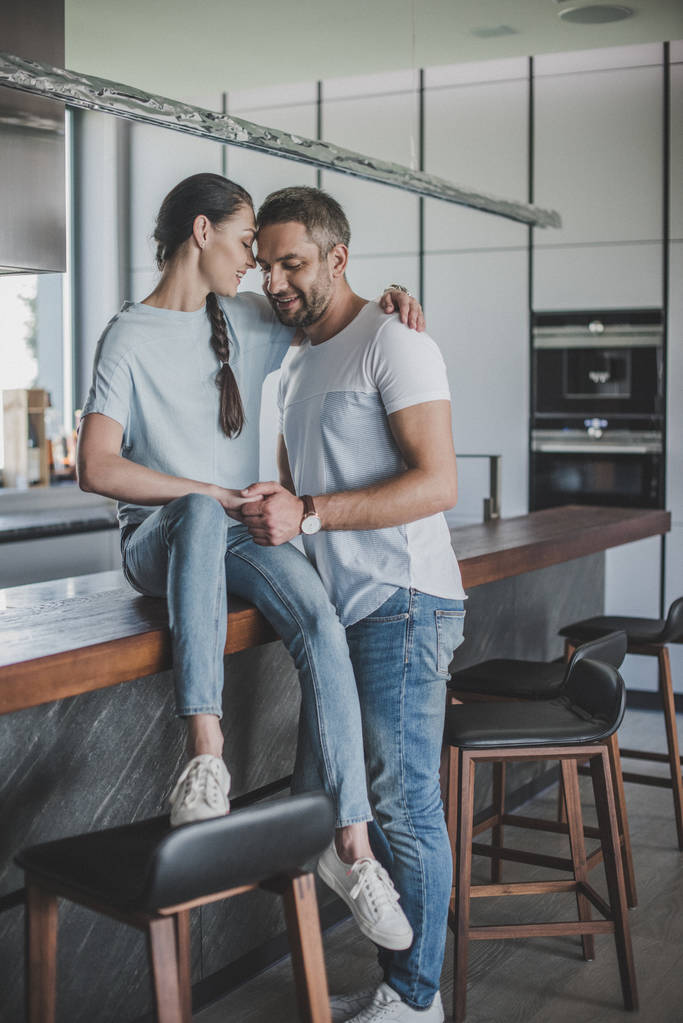 smiling woman sitting on kitchen counter and embracing boyfriend at home - Photo, Image