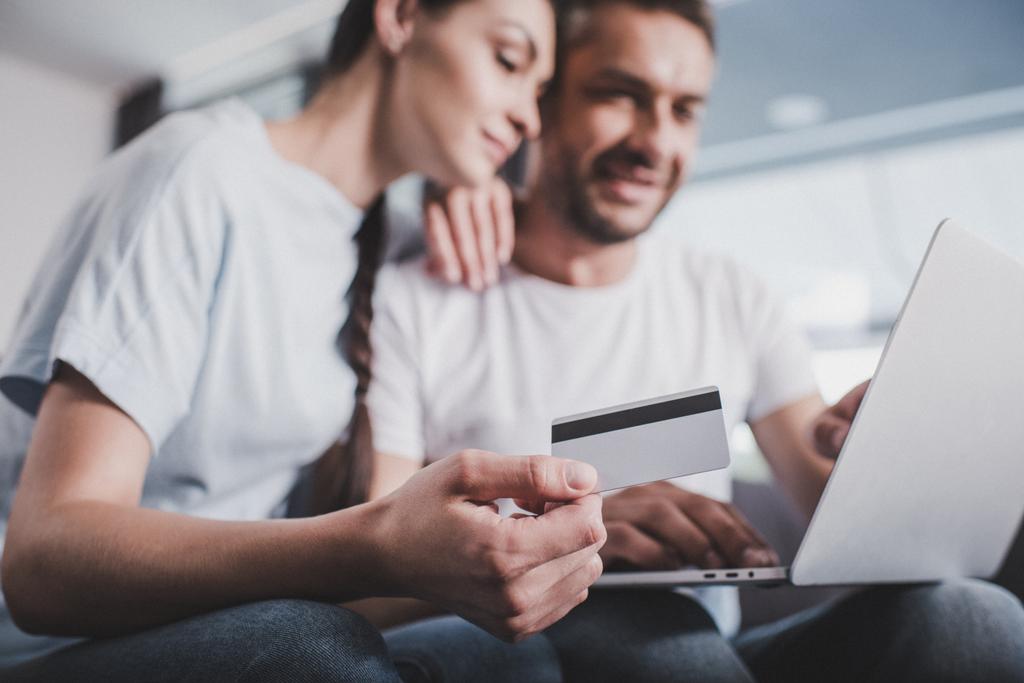 Çift evde kredi kartı ile online alışveriş yaparken dizüstü bilgisayar ile gülümseyen seçici odak - Fotoğraf, Görsel