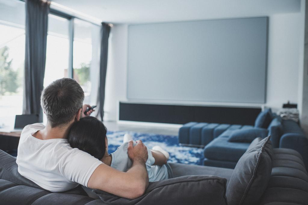 rear view of adult man with remote controller embracing girlfriend and watching tv on sofa at home - Photo, Image