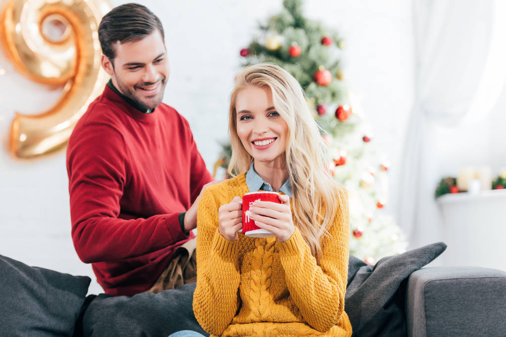 kaunis onnellinen nainen tilalla kahvikuppi kun mies katselee häntä kotona joulukuusi
 - Valokuva, kuva
