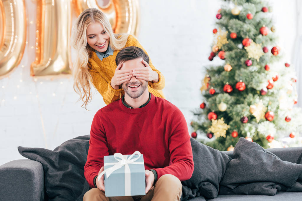 Mädchen schließt Augen und überrascht mit Geschenkbox für Freund zu Hause mit Weihnachtsbaum  - Foto, Bild