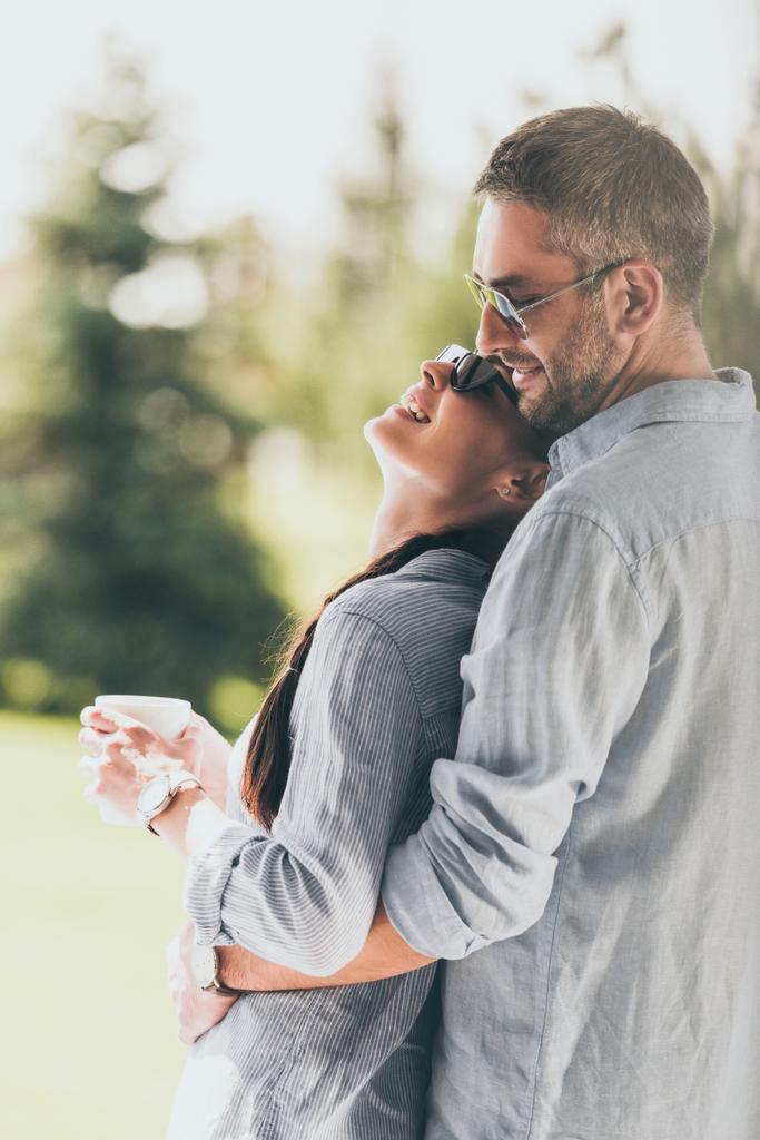 vista lateral de hombre feliz en gafas de sol abrazando novia con taza de café al aire libre
  - Foto, imagen