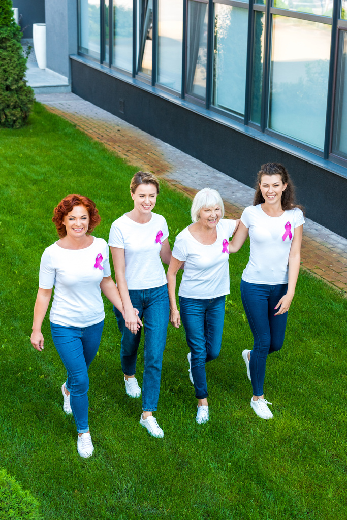 Blick aus der Vogelperspektive auf lächelnde Frauen mit Brustkrebs-Bewusstseinsbändern, die gemeinsam auf grünem Rasen laufen  - Foto, Bild