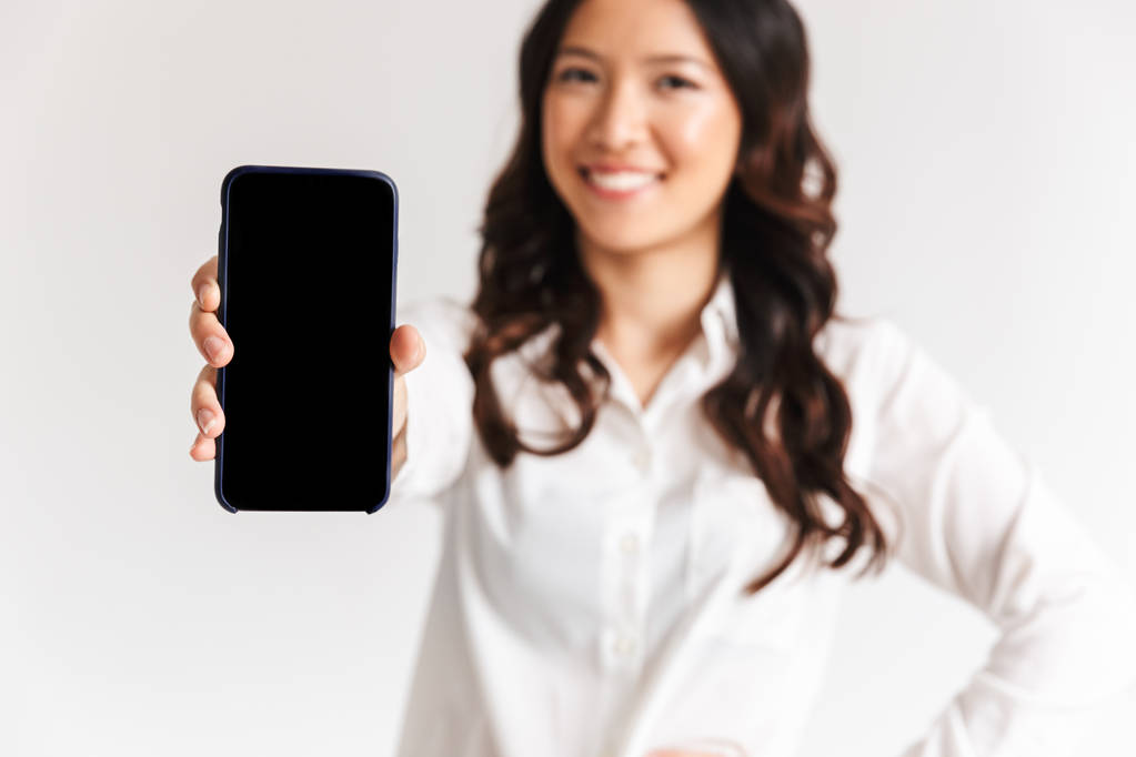 Rozmazane zdjęcie uśmiechający się asian kobieta o długich ciemnych włosach, stałego i pokazywanie smartfona aparat na białym tle nad białym tle w studio - Zdjęcie, obraz