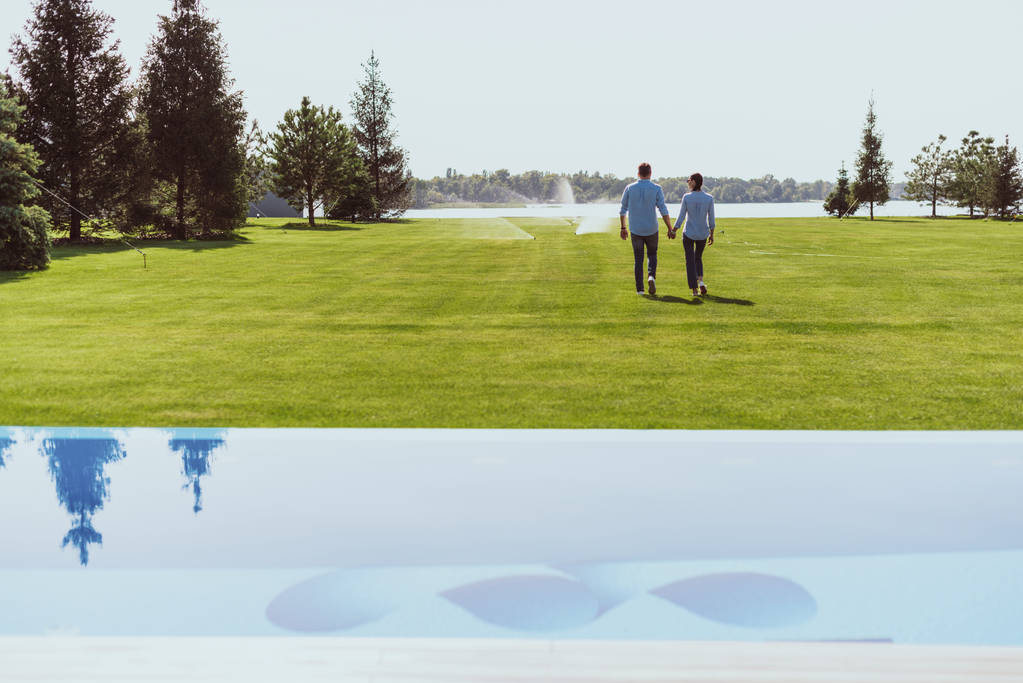 visão traseira do casal de mãos dadas e andando no gramado verde perto da piscina na casa de campo
  - Foto, Imagem