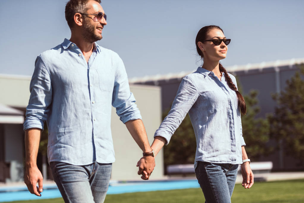 elegante pareja en gafas de sol cogidas de la mano y caminando sobre césped verde en casa de campo
  - Foto, imagen