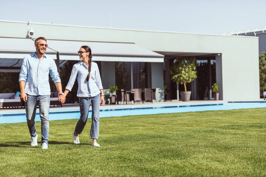 усміхнена пара тримає руки і ходить на зеленому газоні біля басейну в заміському будинку
  - Фото, зображення