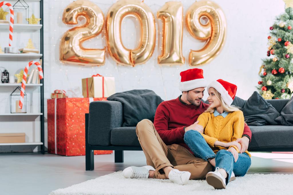 ευτυχισμένο ζευγάρι σε καπέλα santa αγκάλιασμα και κάθεται στο πάτωμα στο σπίτι με μπαλόνια 2019 νέο έτος Χρυσή - Φωτογραφία, εικόνα