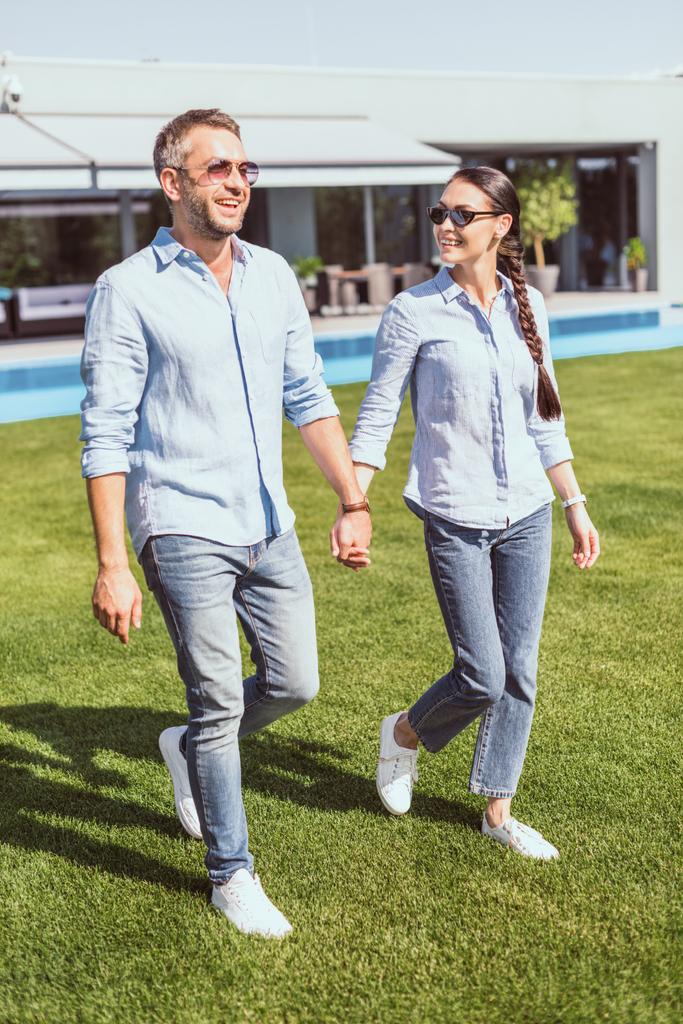happy couple in sunglasses holding hands and walking on green lawn at country house  - Photo, Image