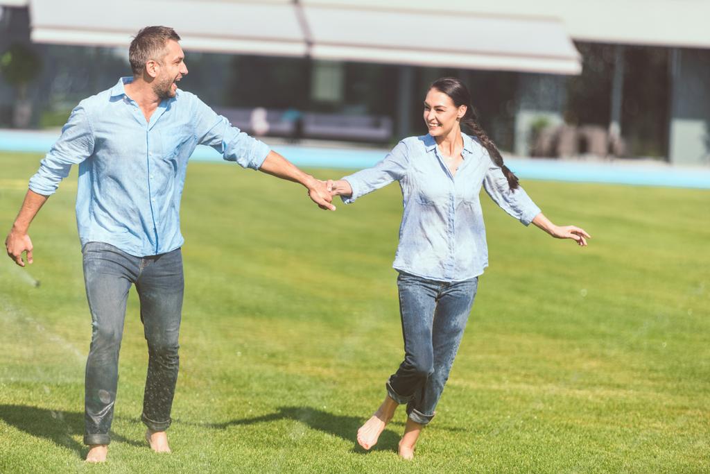 casal sorrindo de mãos dadas e se divertindo com rega automática no gramado verde perto da casa de campo
  - Foto, Imagem