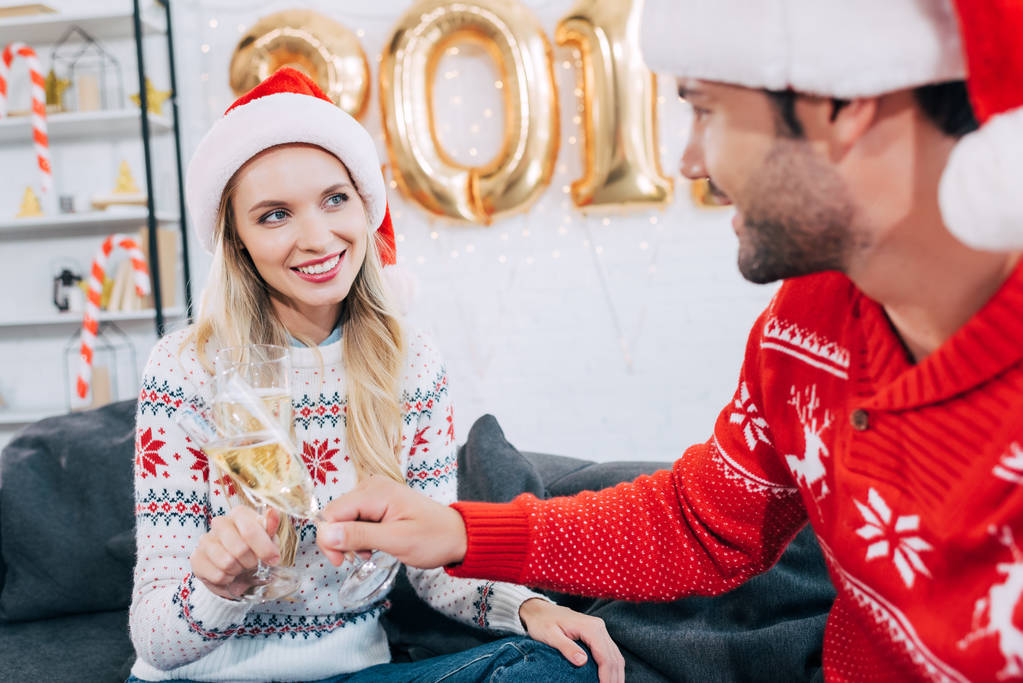 smiling couple in santa hats toasting with champagne glasses and celebrating new year at home - Photo, Image