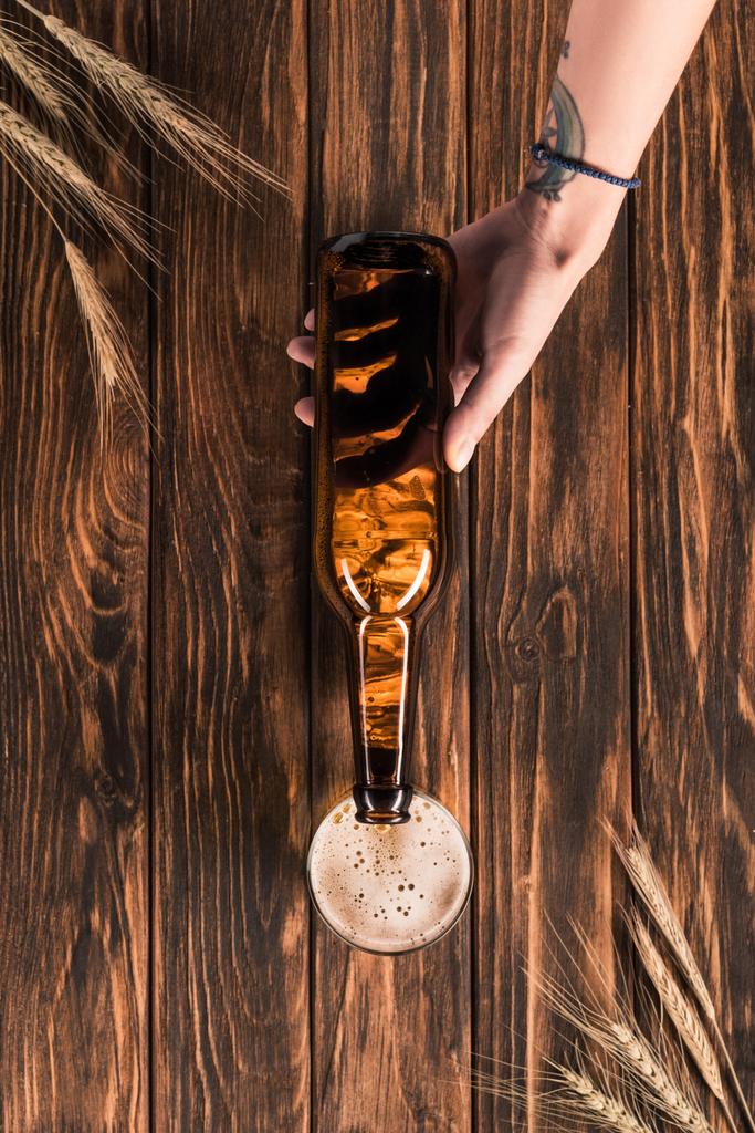 περικομμένη εικόνα γυναίκα ρίχνει μπύρας σε κούπα στο ξύλινο τραπέζι με σιτάρι  - Φωτογραφία, εικόνα