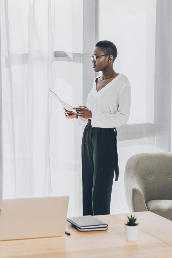 стильная привлекательная африканская американская бизнесвумен, стоящая и читающая документы в офисе
 - Фото, изображение