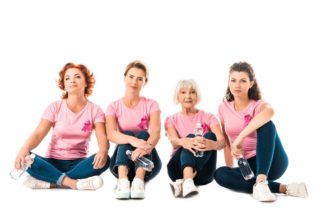 women in pink t-shirts with breast cancer awareness ribbons holding bottles of water and looking at camera while sitting isolated on white - Photo, Image