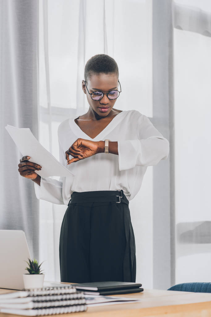 elegante atraente mulher de negócios afro-americana segurando documentos e verificando o tempo no escritório
 - Foto, Imagem