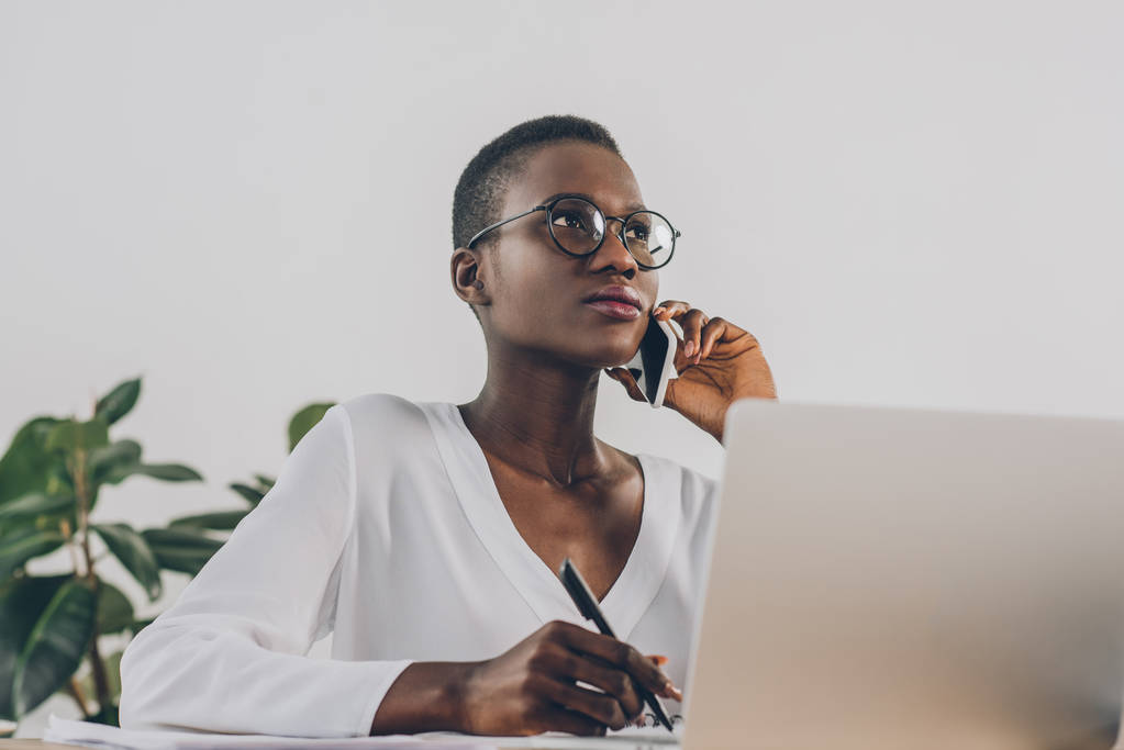 elegante atraente mulher de negócios afro-americana falando por smartphone e olhando para longe no escritório
 - Foto, Imagem