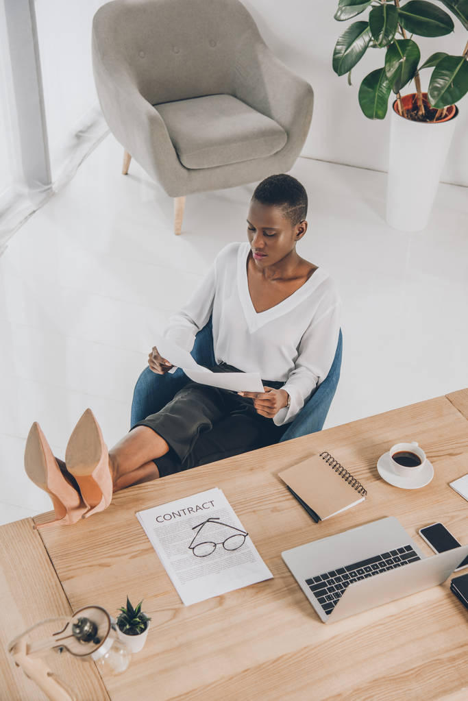 vista de ángulo alto de elegante atractiva mujer de negocios afroamericana lectura de documentos con patas en la mesa en la oficina
 - Foto, Imagen