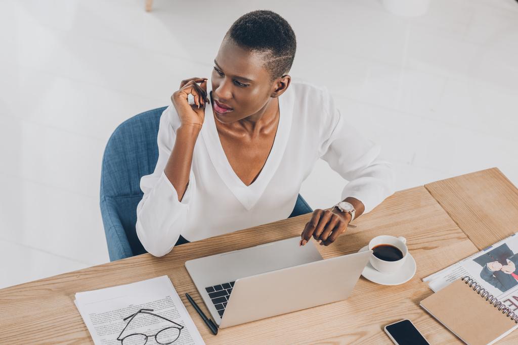 высокий угол зрения стильный привлекательный африканский американский бизнесмен сидя рядом с ноутбуком и глядя в сторону в офисе
 - Фото, изображение