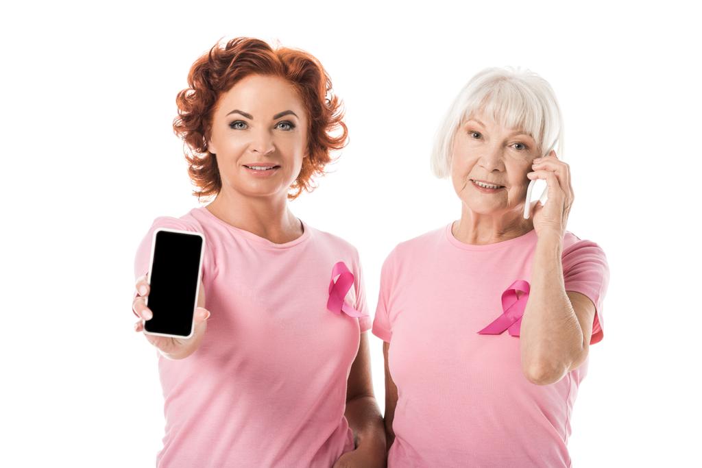 mujeres con cintas de color rosa utilizando teléfonos inteligentes y sonriendo a la cámara aislada en blanco, concepto de conciencia de cáncer de mama
 - Foto, Imagen