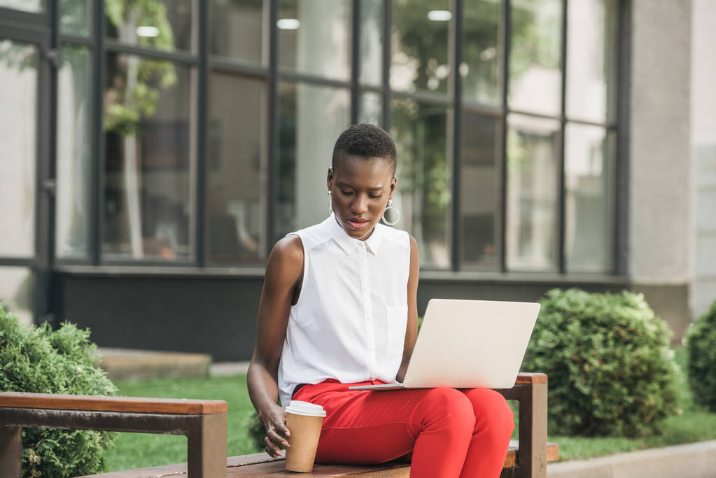 elegante atractiva mujer de negocios afroamericana sentada en el banco con computadora portátil y café para salir a la calle
 - Foto, Imagen