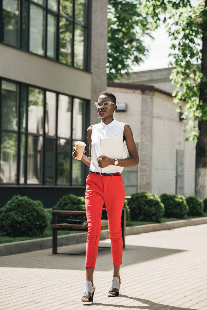 şık çekici Afro-Amerikan işkadını kahve kağıt bardak ve dizüstü bilgisayar ile sokakta yürürken - Fotoğraf, Görsel