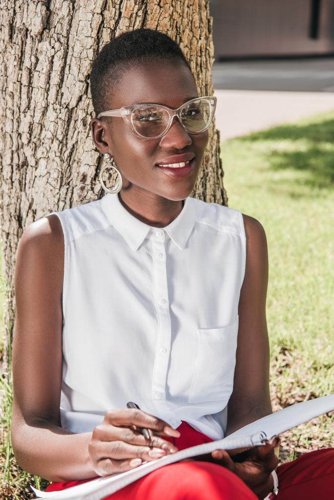elegante y sonriente mujer de negocios afroamericana apoyada en el árbol en el parque y sosteniendo el cuaderno
 - Foto, imagen