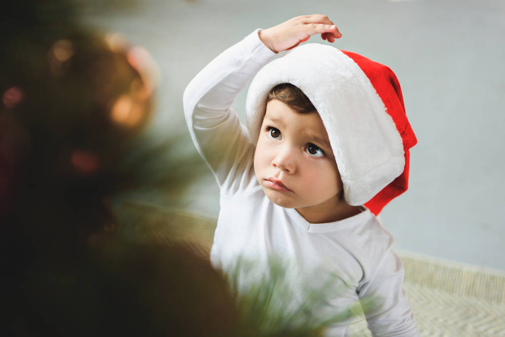 adorable toddler in santa hat looking at christmas tree - Photo, Image