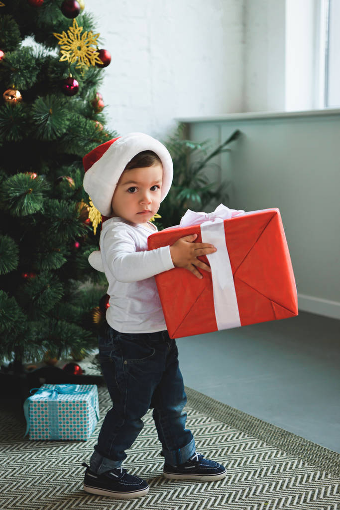 очаровательный ребенок в шляпе Санты с подарками возле рождественской елки
 - Фото, изображение