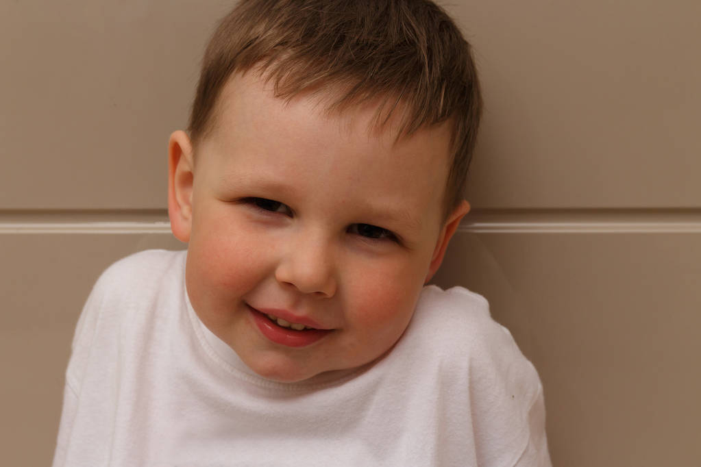 retrato de um menino pequeno, uma criança, em uma camiseta branca, em um fundo bege, olhando para a câmera com desprezo, desconfiança e sarcasmo. tenta sorrir, repugnância, desdém
 - Foto, Imagem