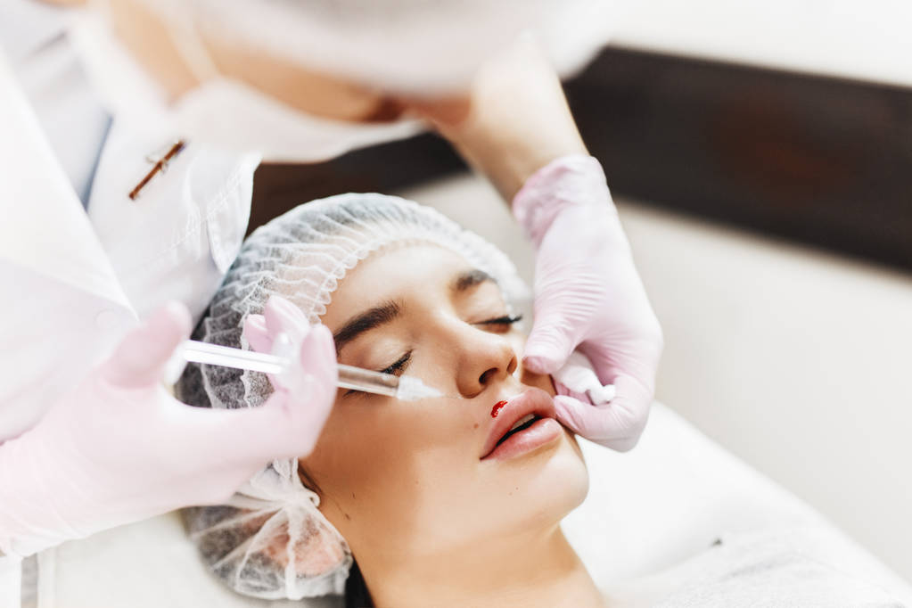 The process of lips enhancement. Cosmetician makes an injection of hyaluronic acid in a beautiful lips. The young girl with a beautiful face in the special hat and hands of the doctor in the pink gloves. - Photo, Image
