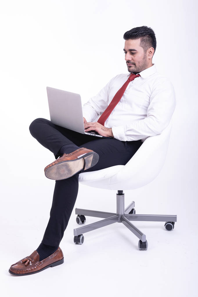 Empresario sentado en silla blanca aislado. Guapo joven empresario indio usando retrato portátil, miradas confiadas. Largura completa tiro lateral
. - Foto, Imagen