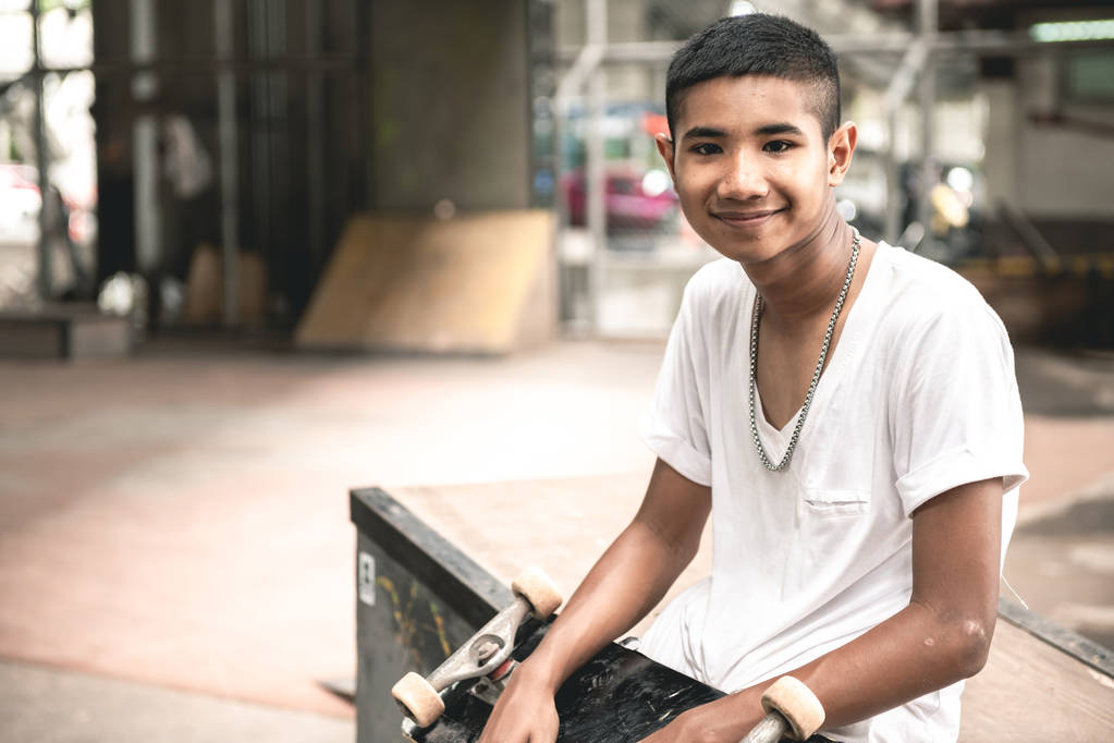 Retrato de skater. Sonriente chico asiático en el parque de skate urbano bajo el puente sentado en hormigón sucio. Entorno de luz natural
. - Foto, Imagen