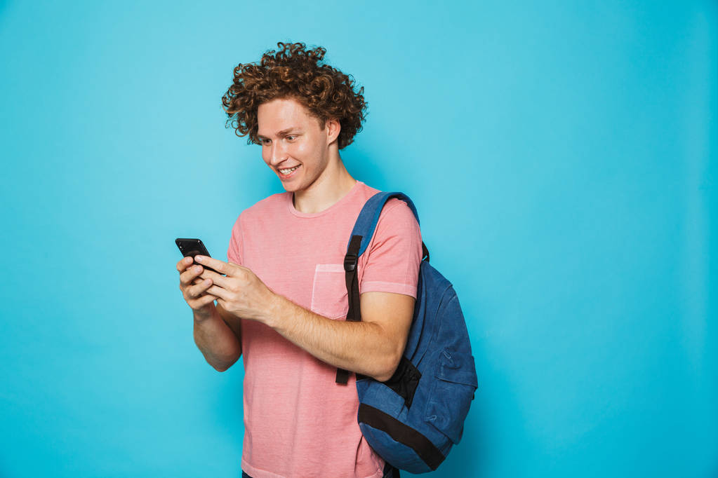 Foto de tipo hipster caucásico con el pelo rizado usando ropa casual y mochila sonriendo y sosteniendo teléfono inteligente aislado sobre fondo azul
 - Foto, imagen