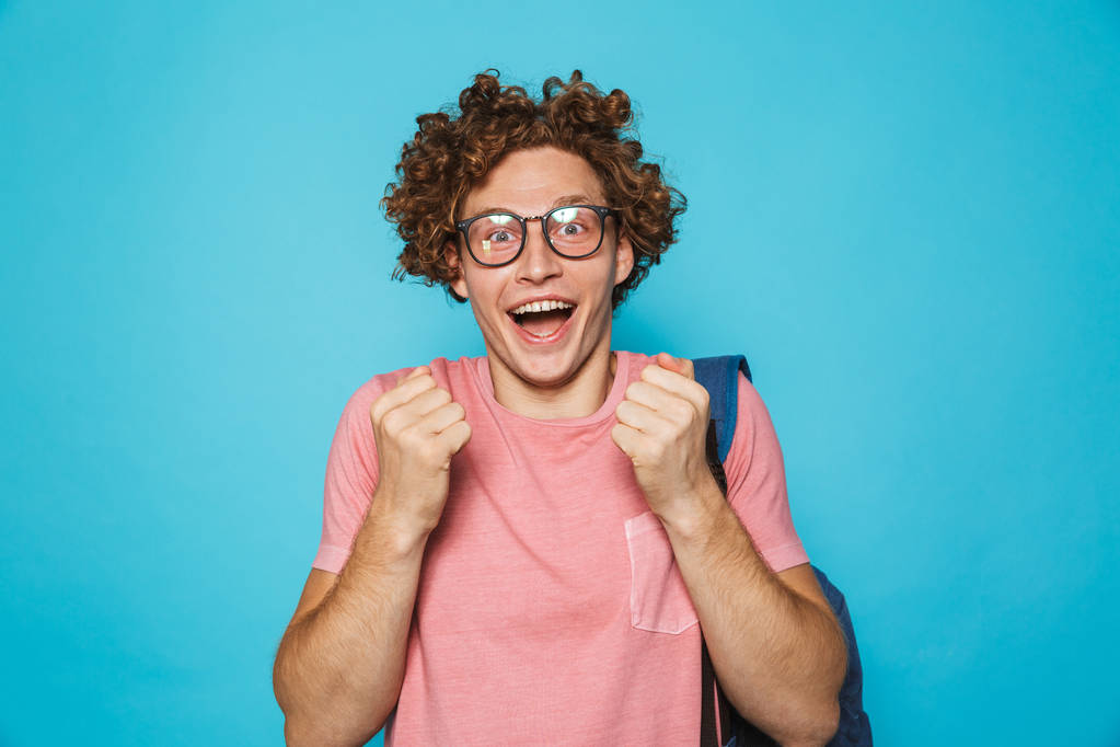 Imagen de tipo con gafas excitadas con pelo rizado usando gafas y mochila sonriendo a la cámara aislada sobre fondo azul
 - Foto, imagen