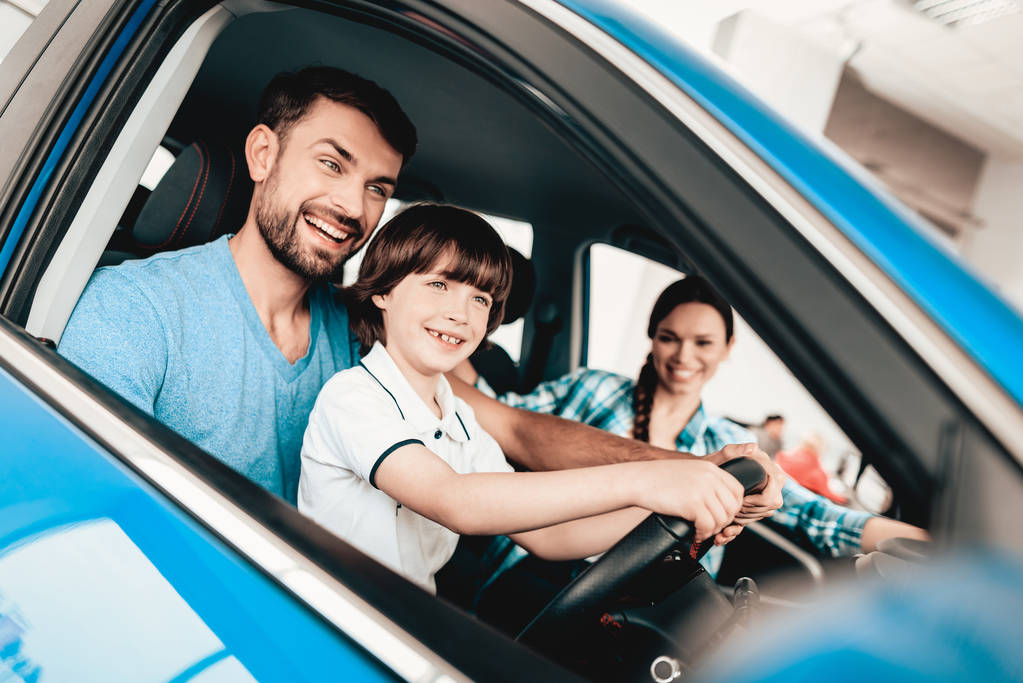 Man With Son Are Sitting At The Wheel Of New Vehicle. Smiling Family. Car Buying In A Showroom. Automobile Salon. Cheerful Driver. Happy Together. Successful Driving. Good Mood. Great Trade. - Photo, Image