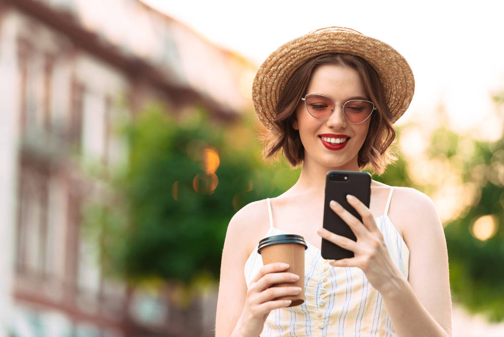 ドレス、麦藁帽子とスマート フォンを屋外で使うサングラスの笑顔の女性のイメージ - 写真・画像