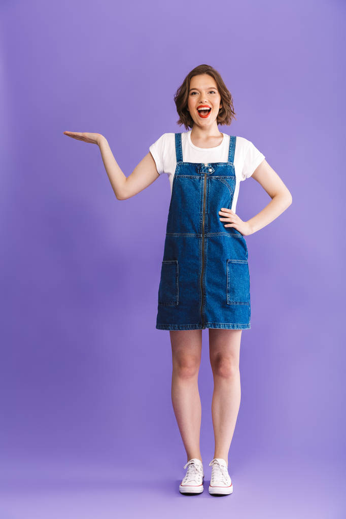 Полнометражный портрет веселой молодой женщины в джинсовой одежде, представляющей пространство для копирования на фиолетовом фоне
 - Фото, изображение