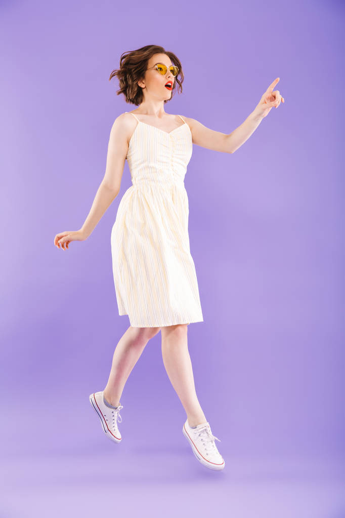 Neşeli bir genç kadının portresi tam uzunlukta ise atlama ve uzağa işaret menekşe arka plan üzerinde poz yaz elbise giymiş - Fotoğraf, Görsel