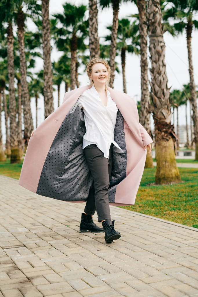 glückliche, stilvolle Frau im langen rosafarbenen Mantel, die im Park spaziert, lächelt und den Frühling genießt - Foto, Bild