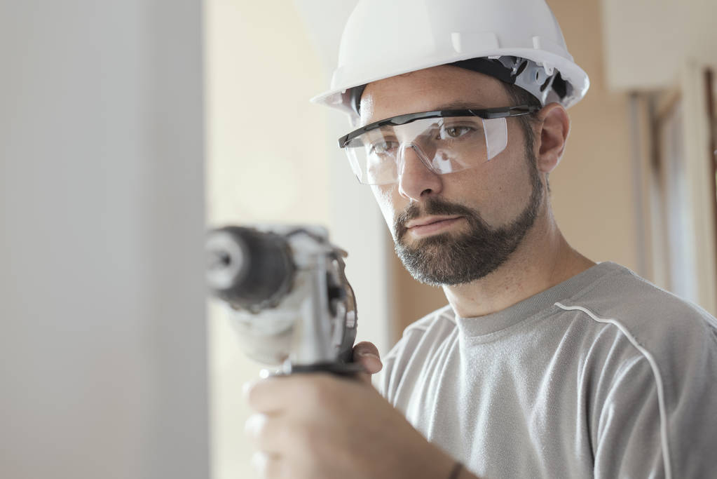 Operaio edile professionista che utilizza un trapano, indossa un casco di sicurezza e occhiali
 - Foto, immagini