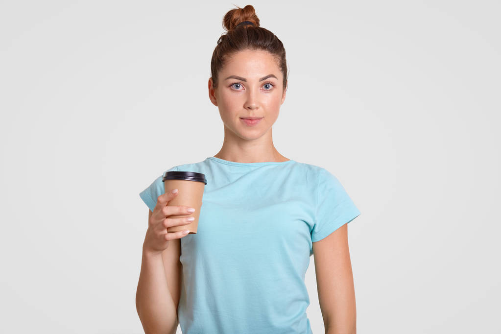 Portrait taille haute d'une femme agréable avec un chignon à cheveux, vêtue d'un t-shirt bleu clair décontracté, tient une tasse de café jetable, a une pause après la gymnastique, a un corps en forme et une peau propre et saine
 - Photo, image