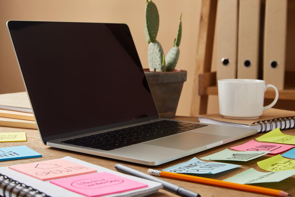 Laptop mit leerem Bildschirm und Papieraufklebern und Kaktus auf der Tischplatte - Foto, Bild