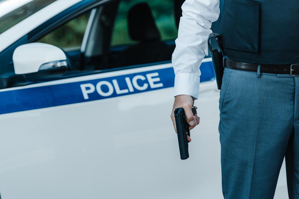 oříznutý obraz policisty v neprůstřelnou vestu, drží zbraň poblíž auto na ulici - Fotografie, Obrázek