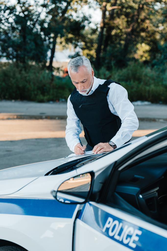 concentrated mature male police officer writing in clipboard on car at street - Photo, Image