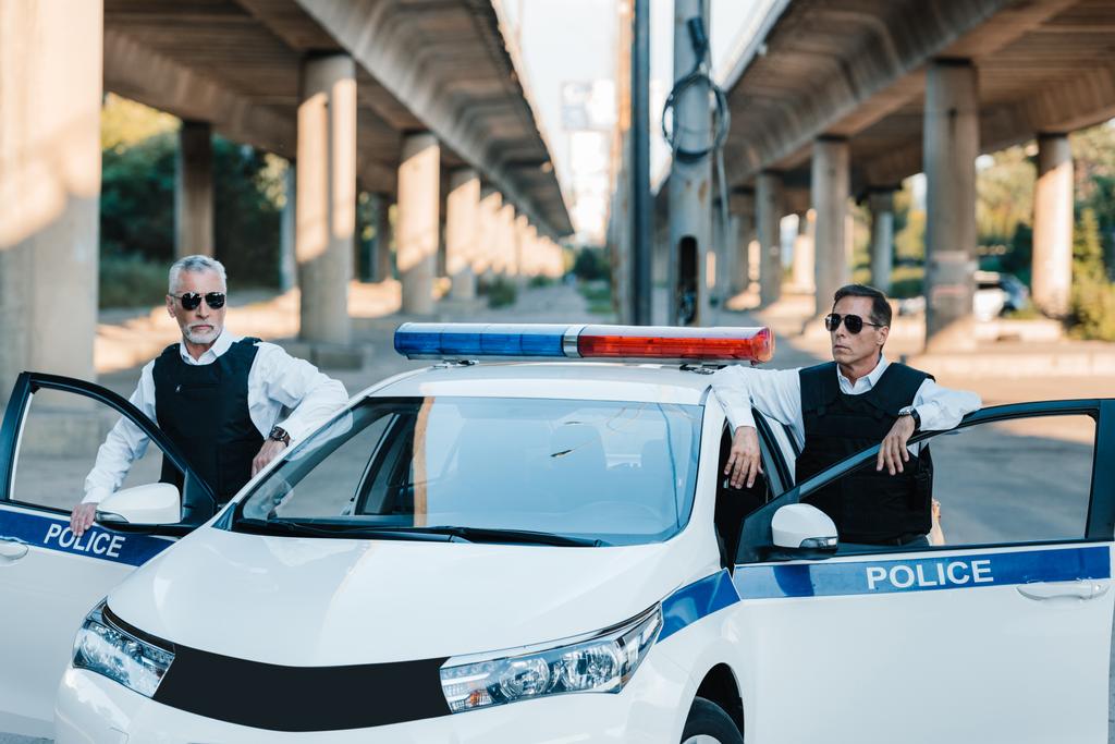 Selbstbewusster reifer Polizist in Sonnenbrille und schusssicherer Weste steht neben Auto an der Stadtstraße  - Foto, Bild