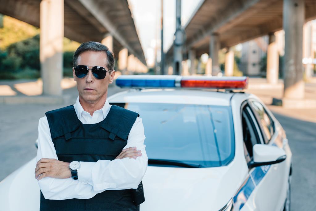 maturo poliziotto in occhiali da sole e giubbotto antiproiettile in piedi con braccia incrociate vicino auto in strada
 - Foto, immagini