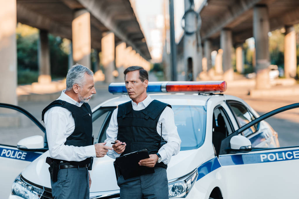 Polizist mittleren Alters spricht mit Kollegin mit Klemmbrett neben Auto auf Straße - Foto, Bild