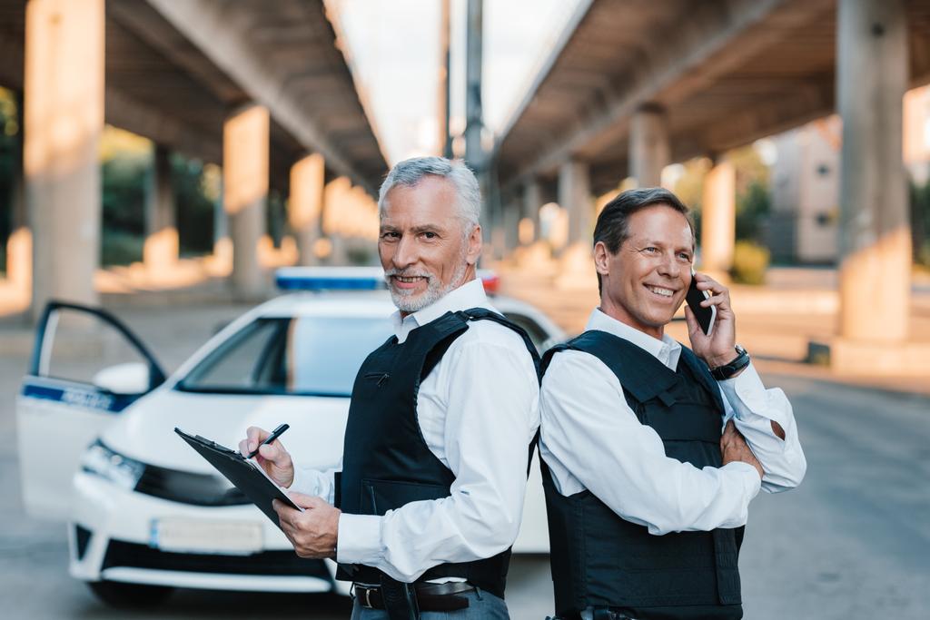 oficial de policía masculino sonriente hablando en el teléfono inteligente mientras su colega escribe en portapapeles en el coche en la calle
 - Foto, imagen