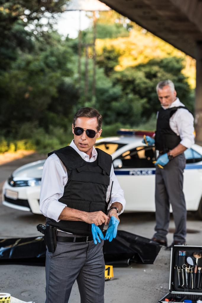 poważne policjanta w okulary zakładanie rękawic lateksowych, podczas gdy jego kolega stojący na zbrodni z trupem w ciało torba  - Zdjęcie, obraz